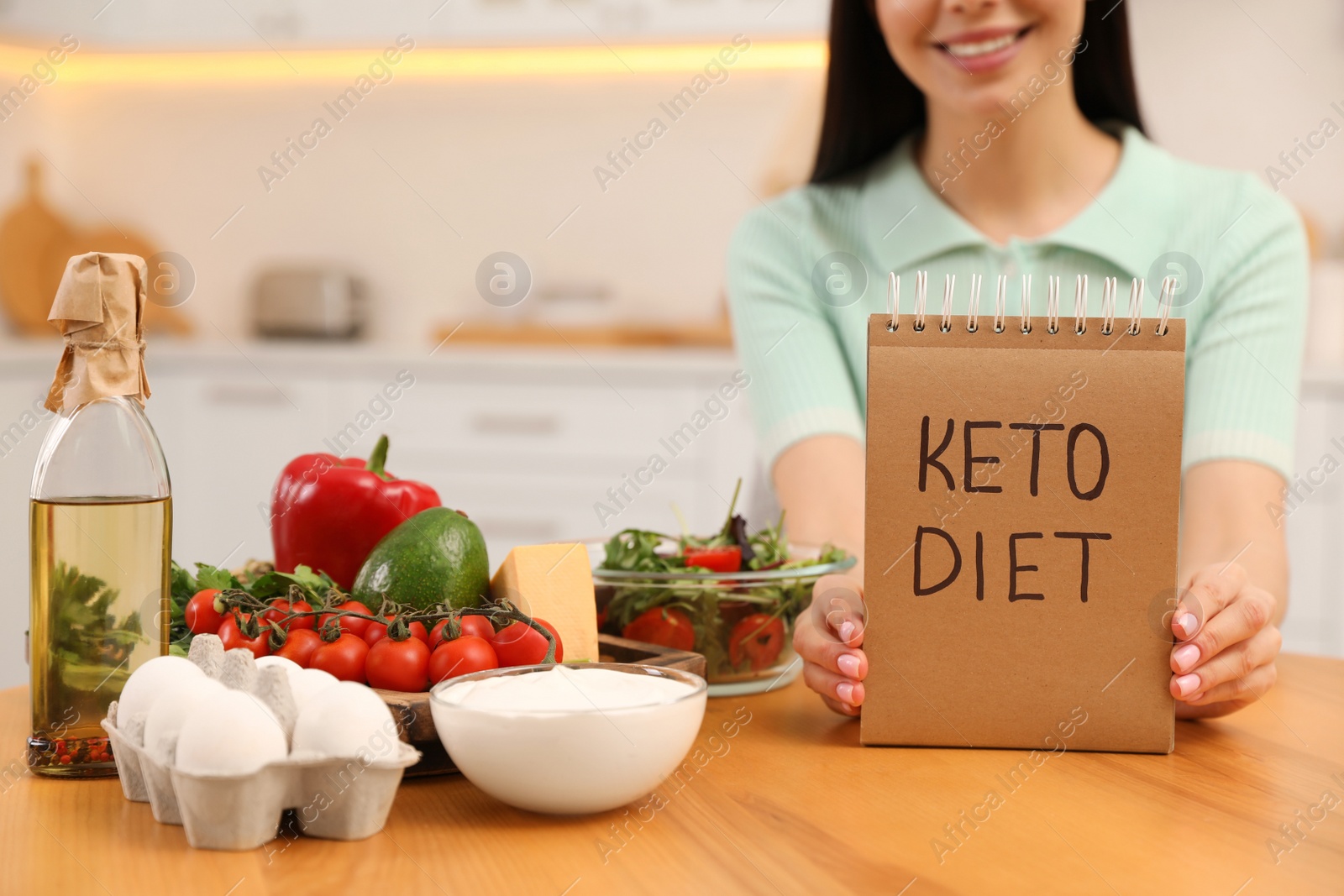 Photo of Happy woman holding notebook with words Keto Diet near different products in kitchen, closeup