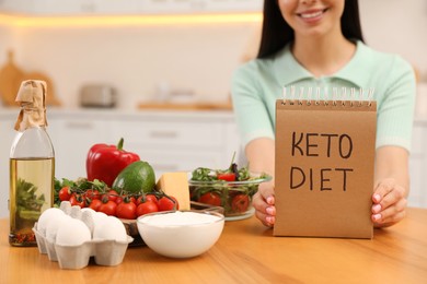 Photo of Happy woman holding notebook with words Keto Diet near different products in kitchen, closeup