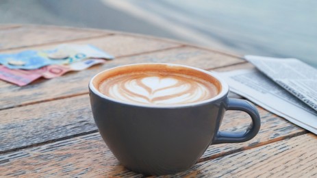 Photo of Cup of delicious coffee, newspaper and banknotes on wooden table, closeup