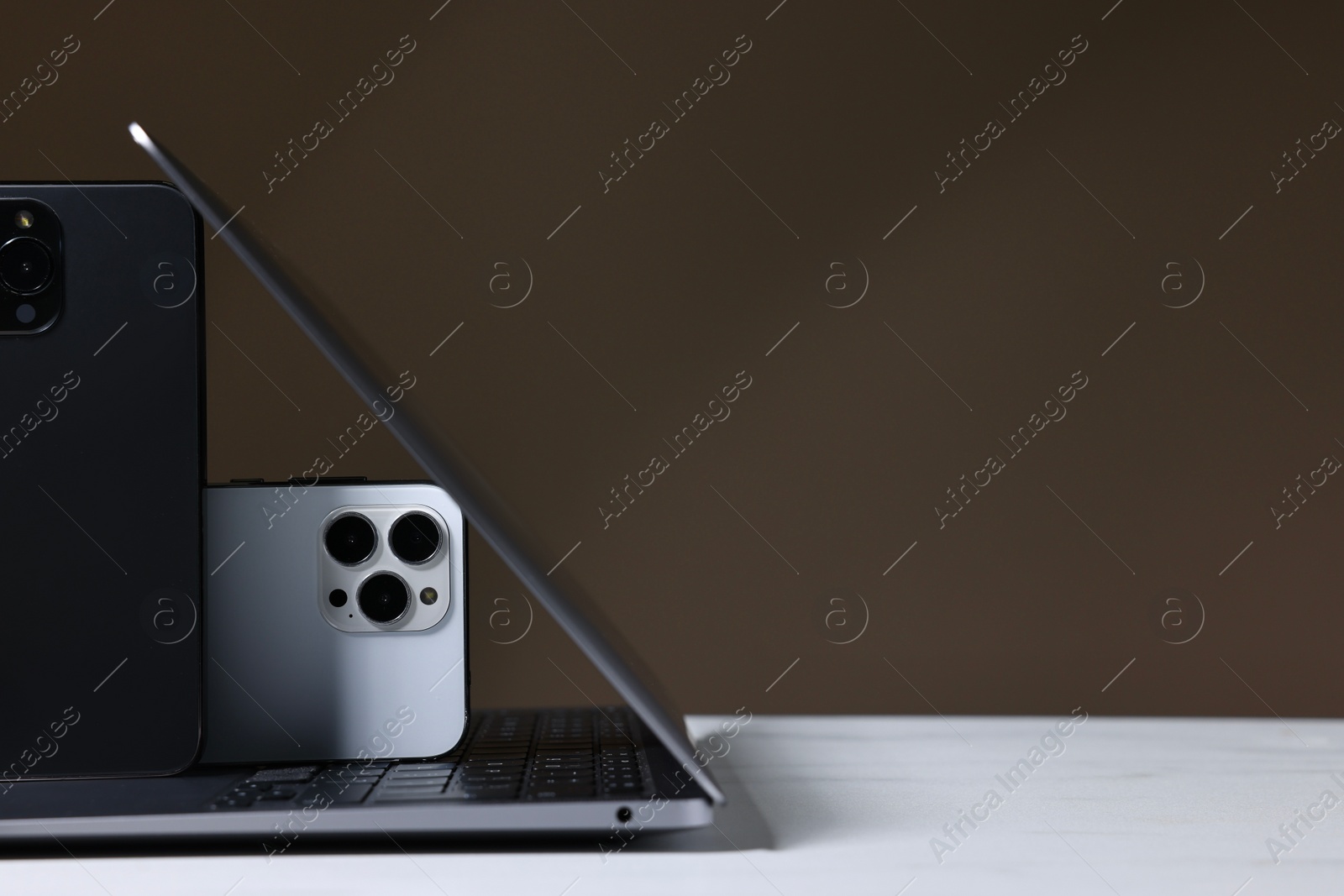 Photo of Modern laptop and smartphones on white table against brown background. Space for text