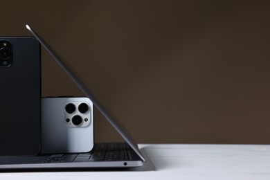 Photo of Modern laptop and smartphones on white table against brown background. Space for text