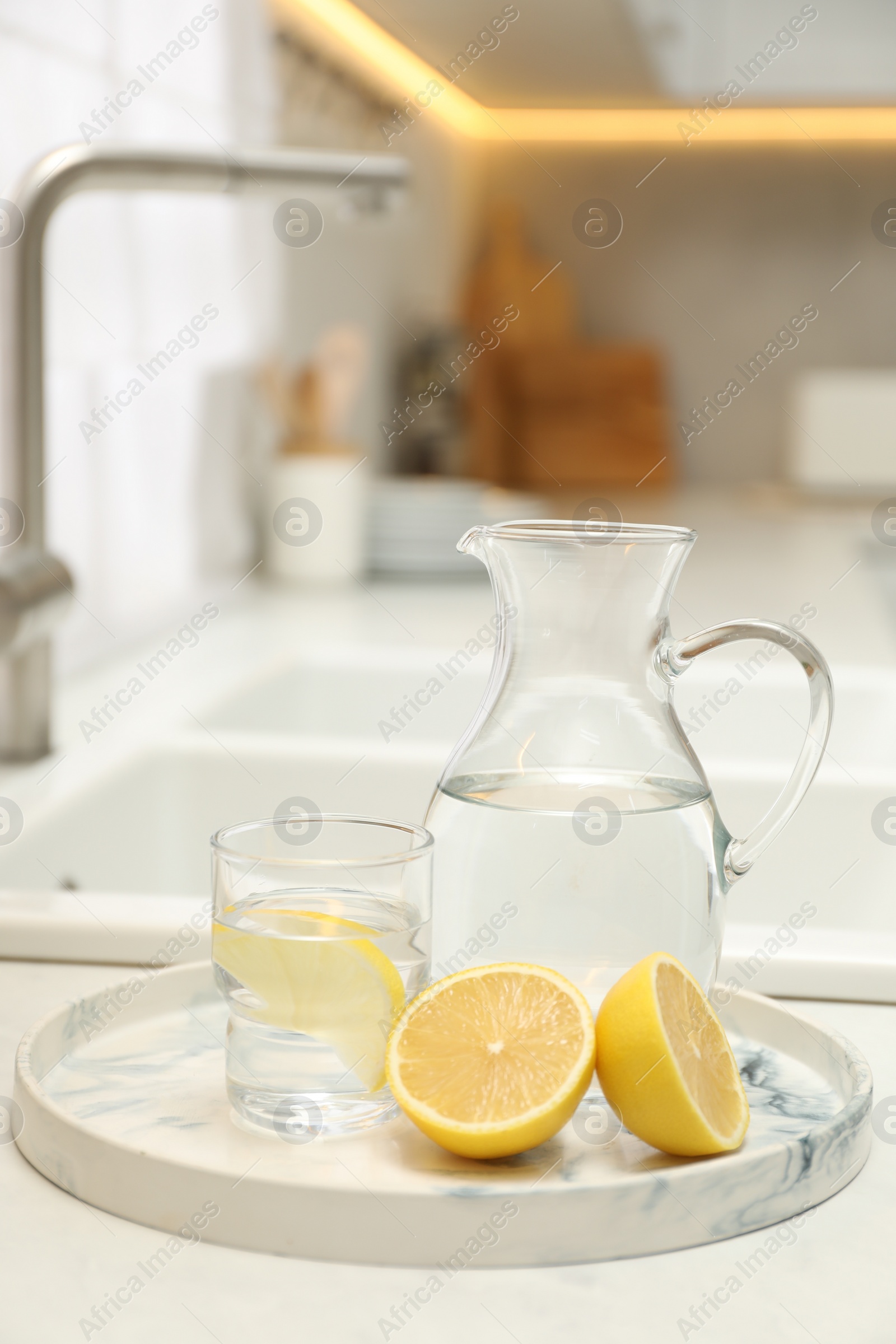 Photo of Jug, glass with clear water and lemons on white table in kitchen