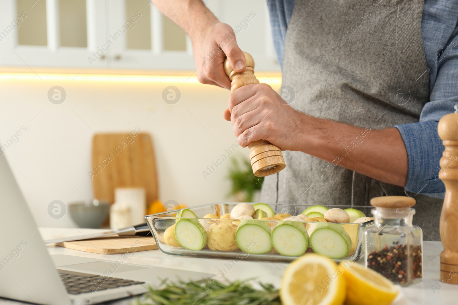 Photo of Man adding spices to dish while watching online cooking course via laptop in kitchen, closeup