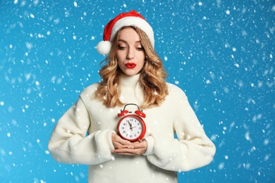 Image of Happy young woman wearing Santa hat with alarm clock on light blue background. Christmas time
