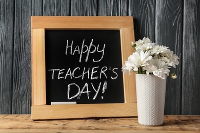 Photo of Chalkboard with inscription HAPPY TEACHER'S DAY and vase of flowers on wooden table against grey wall