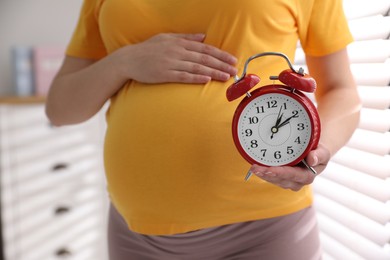 Photo of Young pregnant woman holding alarm clock near her belly at home, closeup. Time to give birth