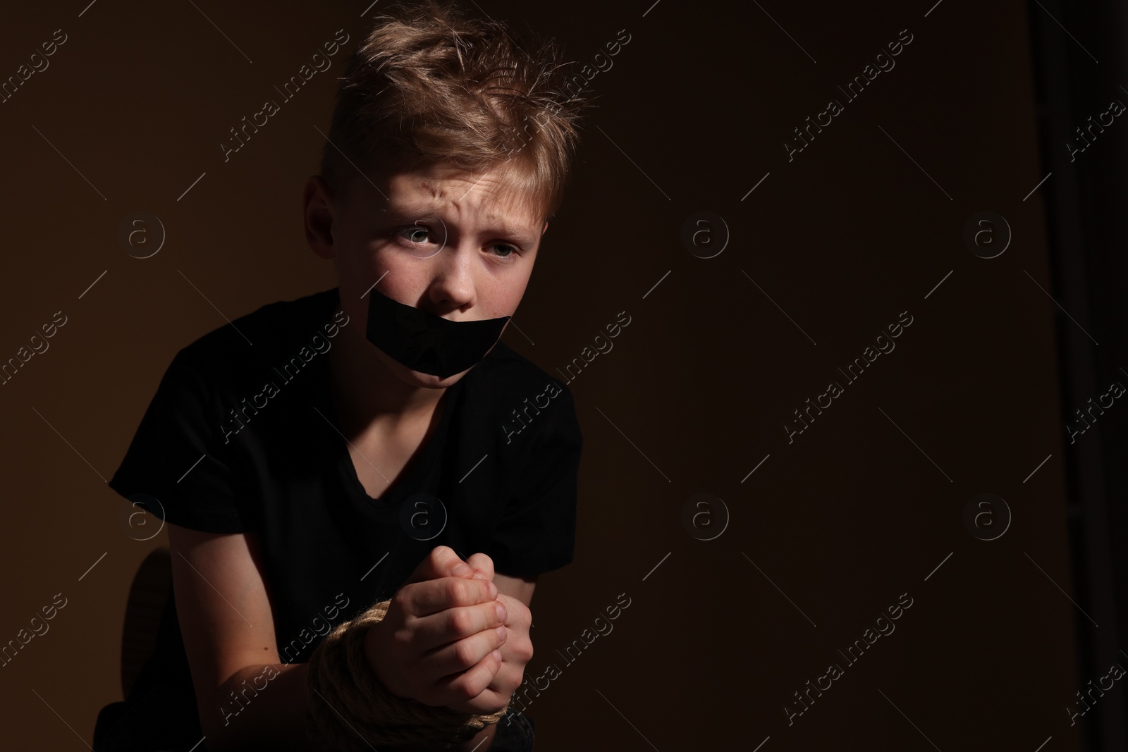 Photo of Little boy with taped mouth tied up and taken hostage against dark background. Space for text