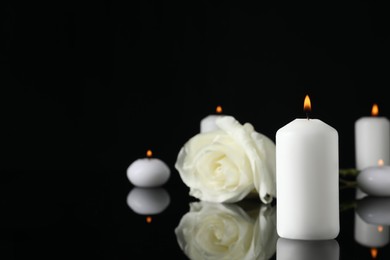Photo of White rose and burning candles on black mirror surface in darkness, space for text. Funeral symbols
