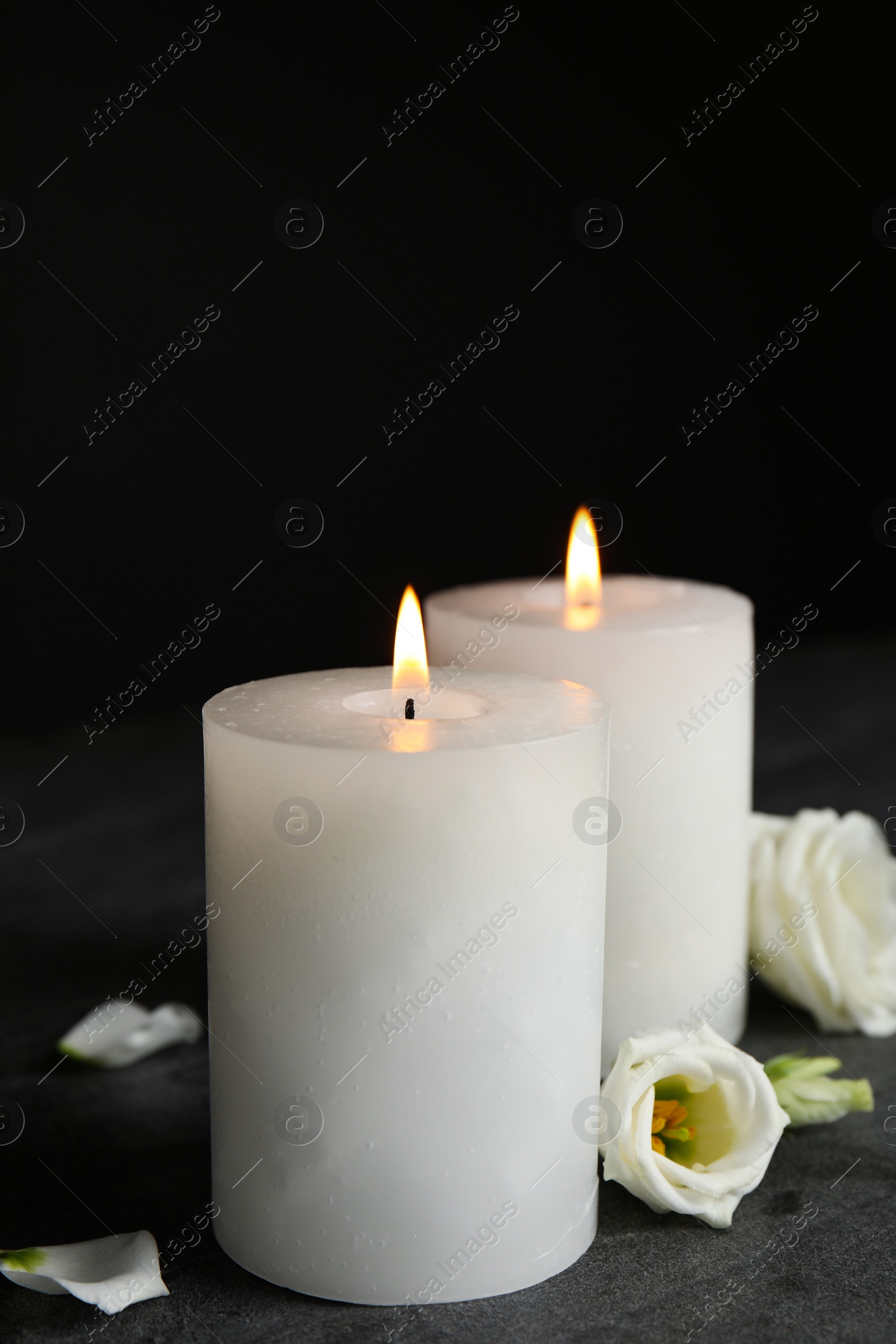 Photo of Burning candles and flowers on grey table against black background, space for text