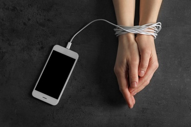 Photo of Woman holding hands tied with charging cable near mobile phone on grey background, above view. Loneliness concept