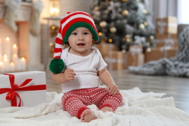 Cute little baby with elf hat near Christmas gift on floor at home