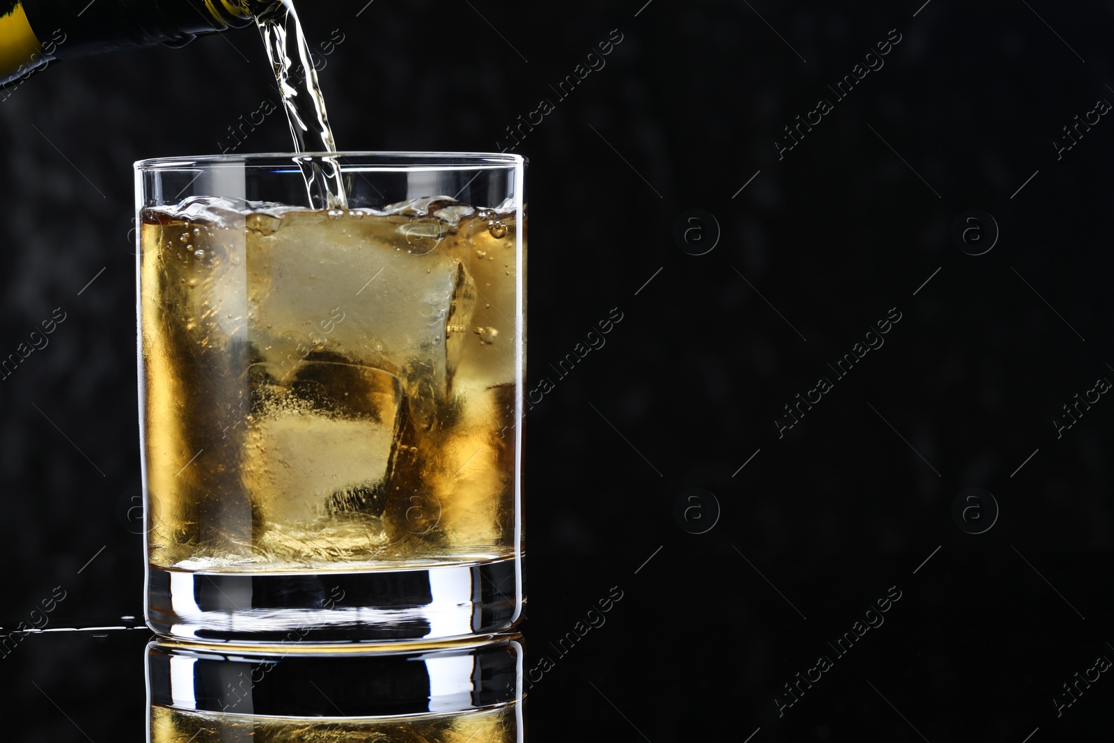 Photo of Pouring tasty whiskey from bottle into glass at mirror table against black background, closeup. Space for text