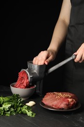 Photo of Woman making beef mince with manual meat grinder at dark textured table against black background, closeup