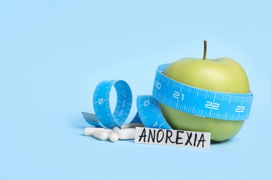 Photo of Paper with word anorexia, apple, measuring tape and pills on light blue background
