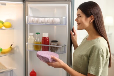 Happy woman holding bowl covered with beeswax food wrap near refrigerator in kitchen