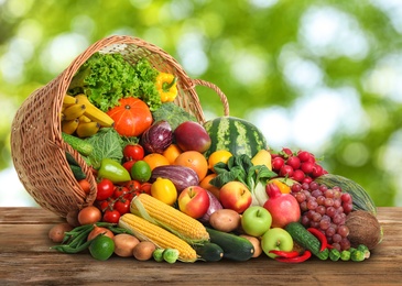 Image of Overturned wicker basket with different fresh organic vegetables and fruits on wooden table against blurred green background