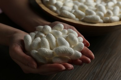 Woman holding white silk cocoons over bowl at wooden table, closeup