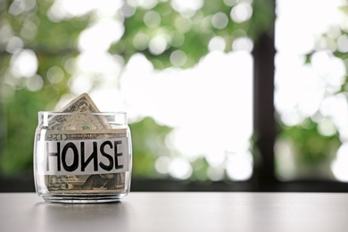 Glass jar with money and word HOUSE on table against blurred background, space for text