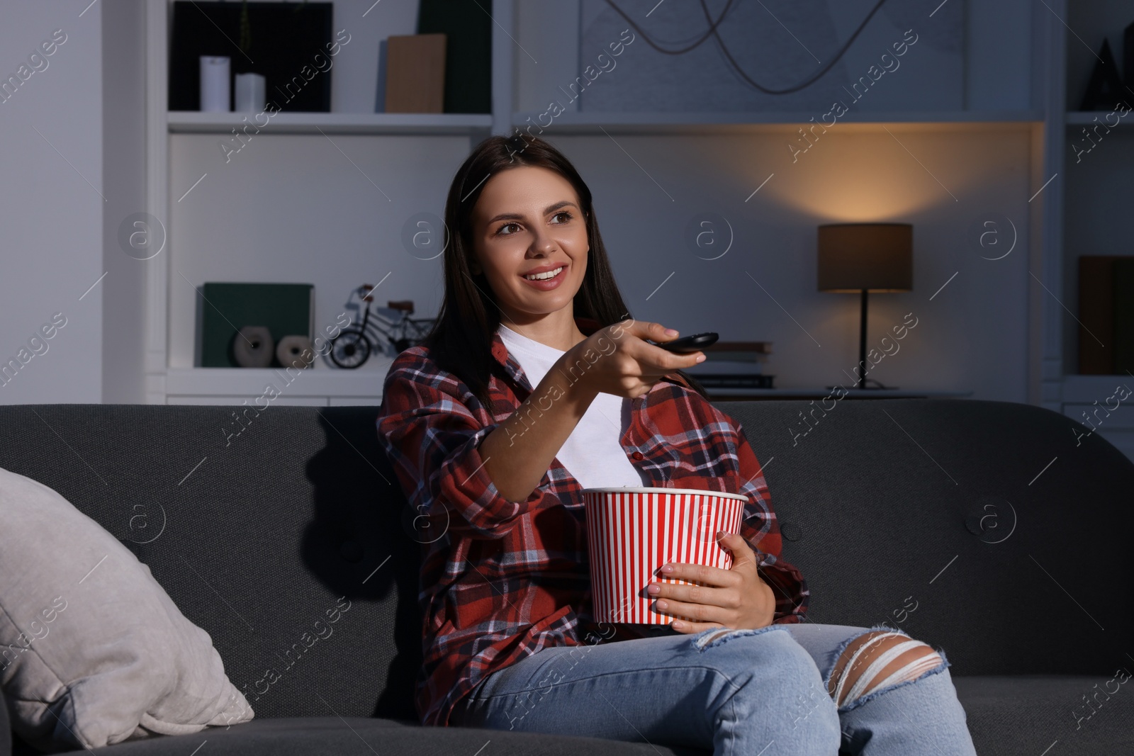 Photo of Happy woman holding popcorn bucket and changing TV channels with remote control at home in evening