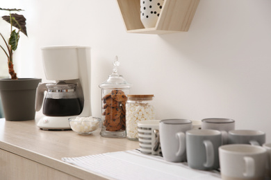 Photo of Modern coffeemaker and cups on commode near light wall indoors