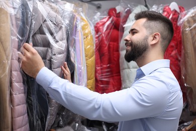 Photo of Dry-cleaning service. Happy worker choosing clothes from rack indoors