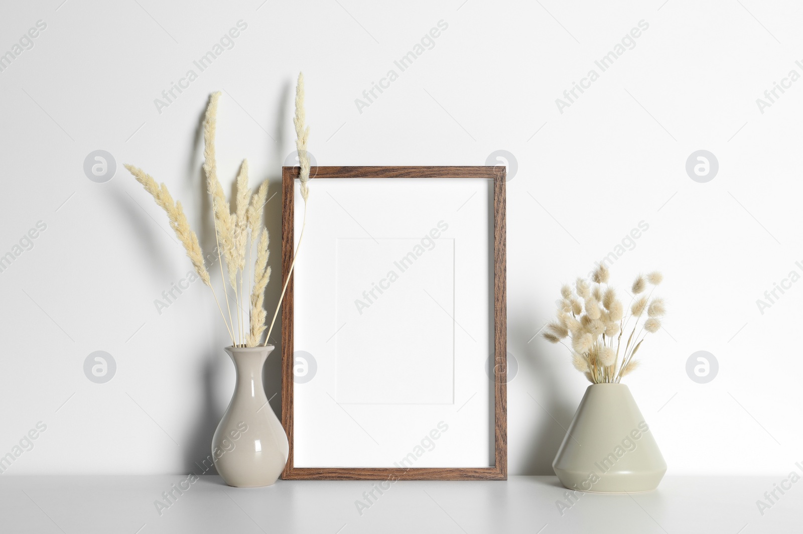 Photo of Empty photo frame and vases with dry decorative spikes on white table. Mockup for design