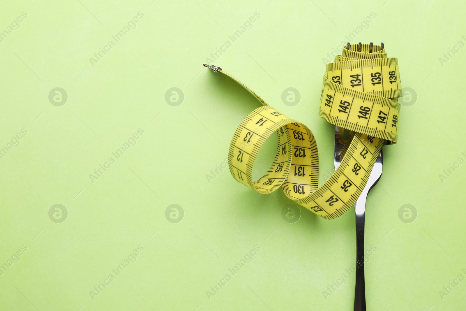 Photo of Fork with measuring tape on light green background, top view and space for text. Diet concept