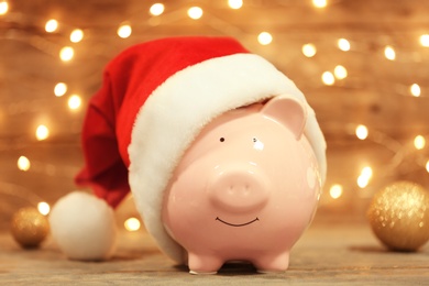 Cute piggy bank with Santa hat on table against blurred Christmas lights