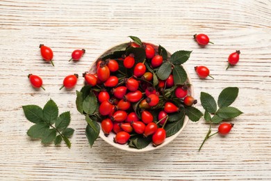Photo of Ripe rose hip berries with green leaves on white wooden table, flat lay