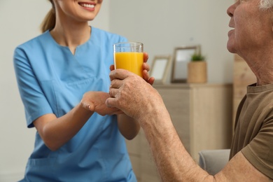 Nurse giving glass of juice to elderly man indoors, closeup. Assisting senior people