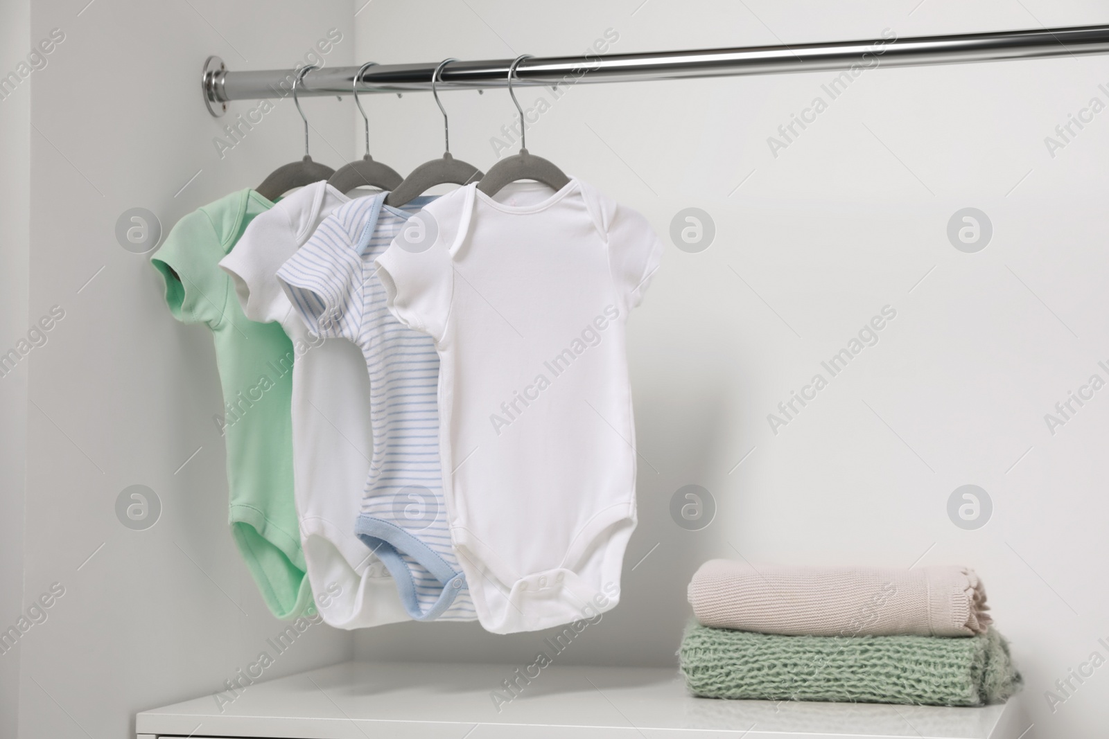 Photo of Hangers with baby bodysuits and stack of clothes near white wall. Space for text