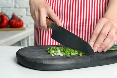 Woman cutting green spring onion on black wooden board at white table, closeup