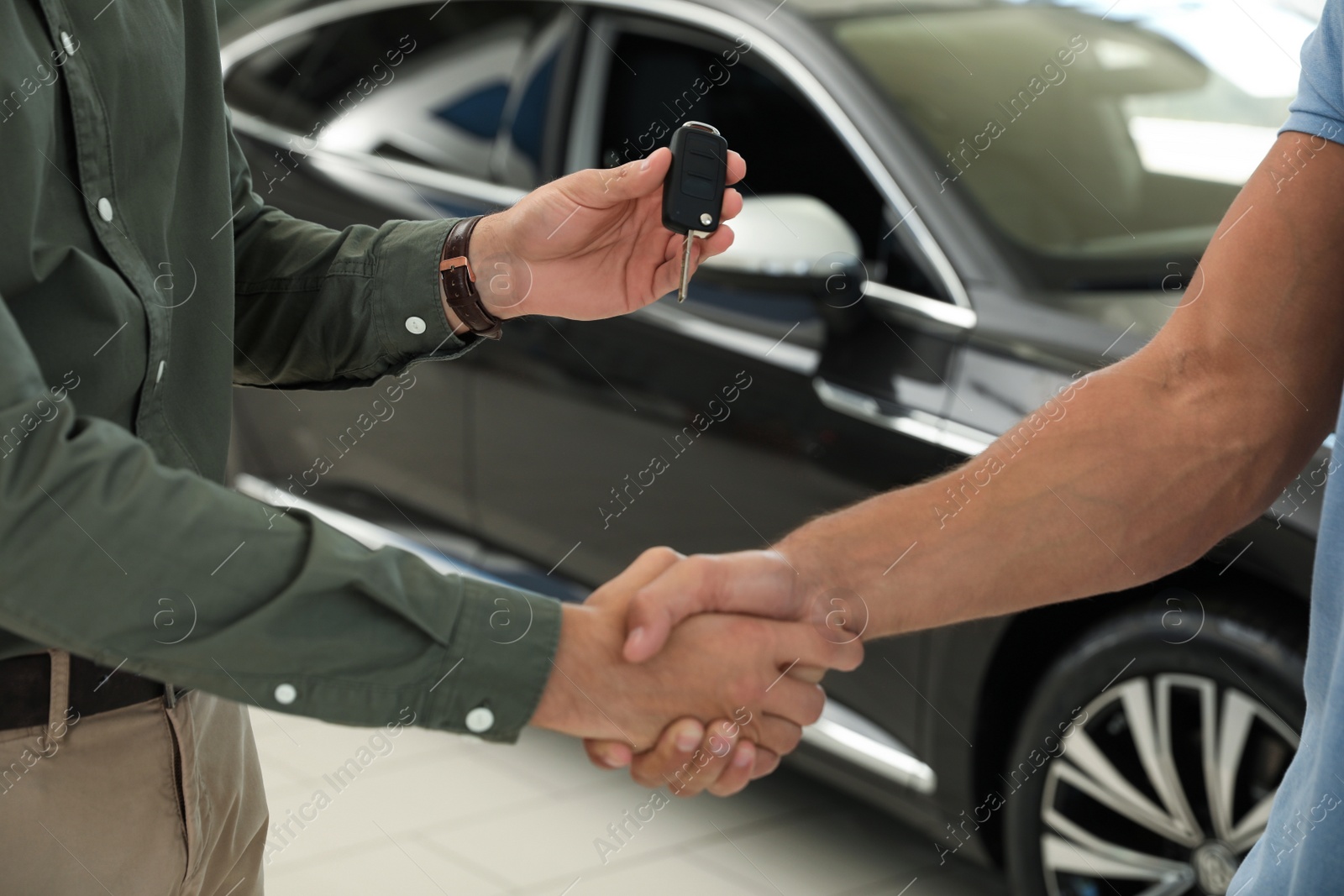 Photo of Young salesman shaking hands with customer and giving car key in modern auto dealership, closeup