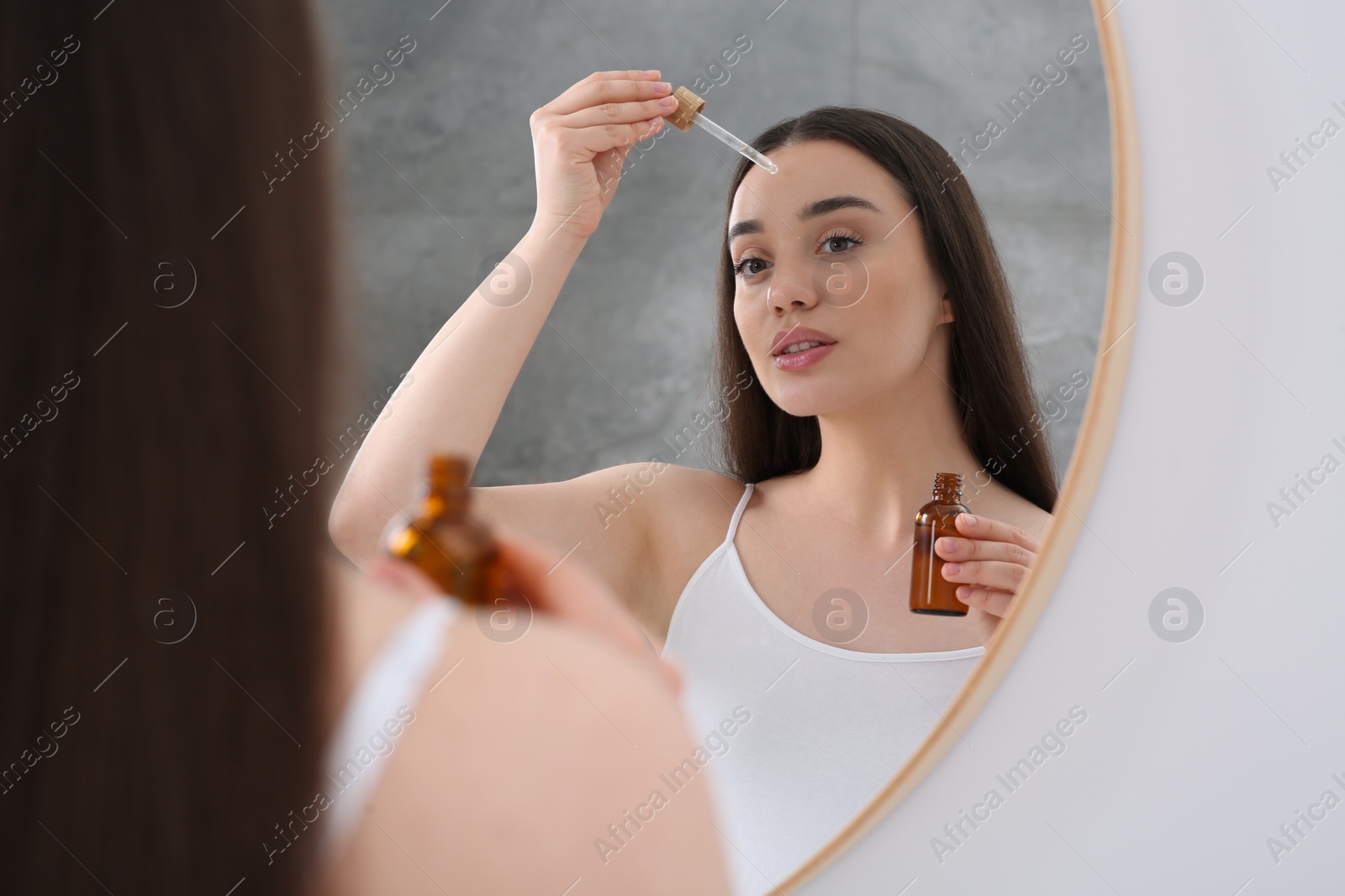 Photo of Woman applying essential oil onto face near mirror