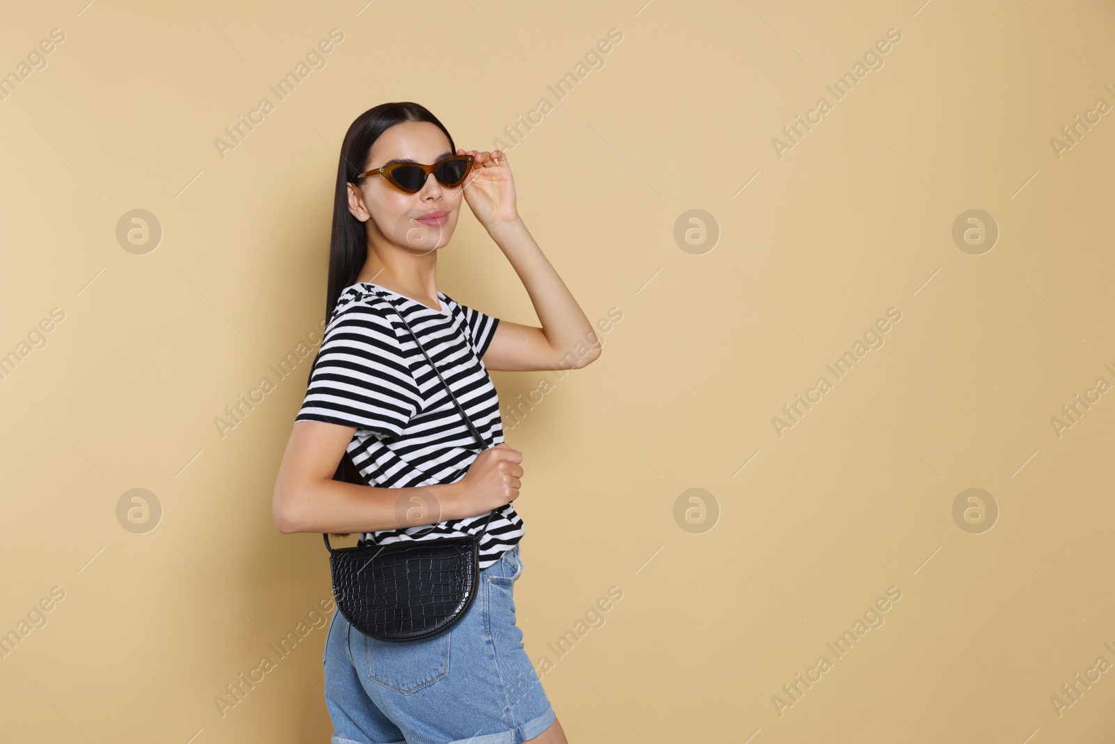 Photo of Beautiful young woman with stylish bag in sunglasses on beige background, space for text