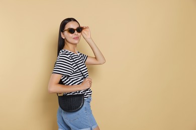 Photo of Beautiful young woman with stylish bag in sunglasses on beige background, space for text
