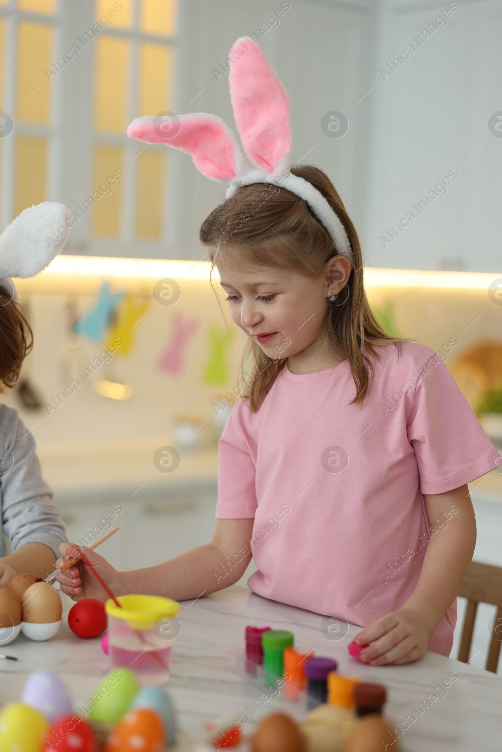 Photo of Easter celebration. Children with bunny ears painting eggs at white marble table in kitchen