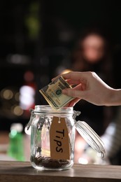 Photo of Woman putting tips into glass jar on wooden table indoors, closeup