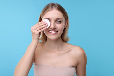 Young woman cleaning face with cotton pad on light blue background