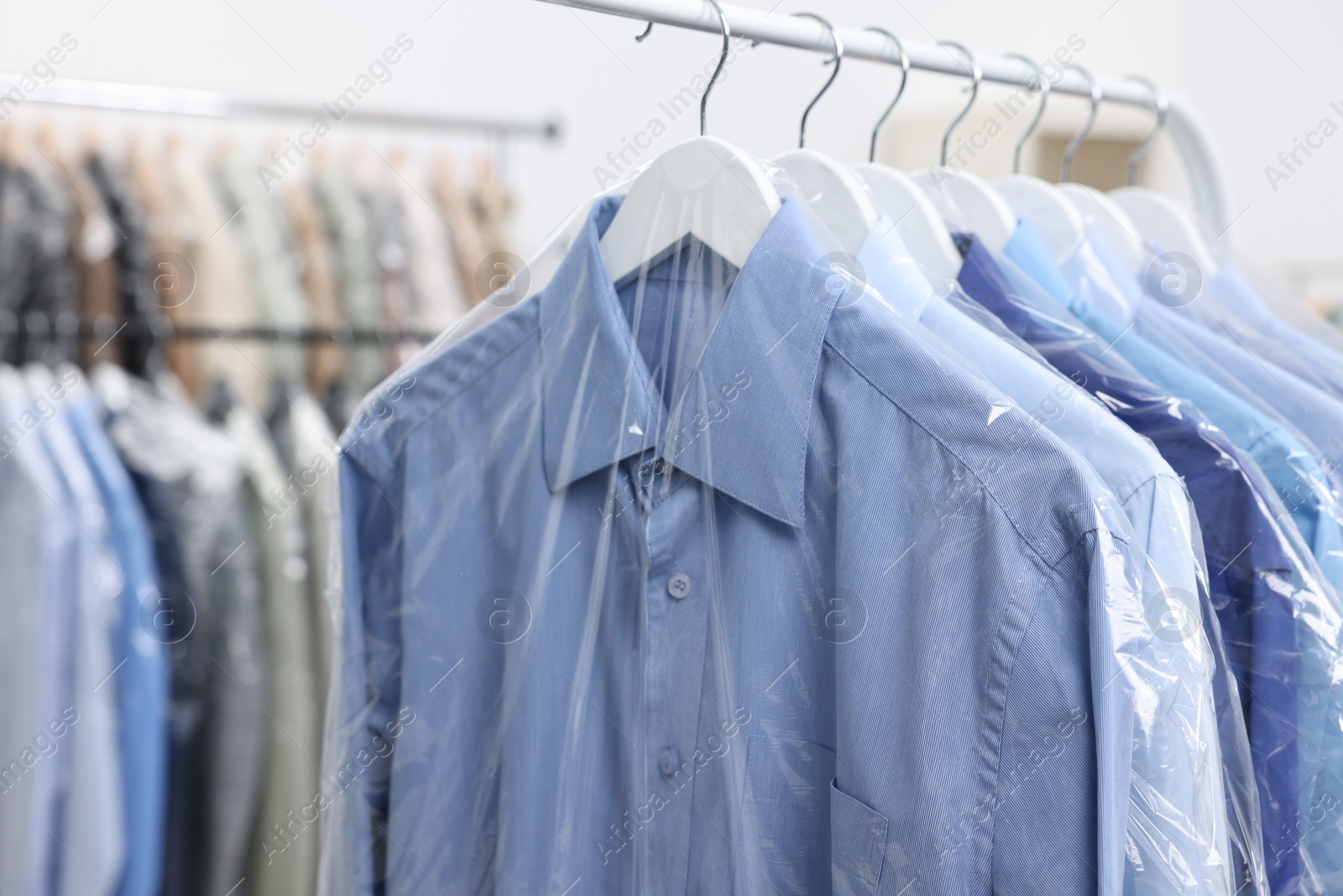 Photo of Dry-cleaning service. Many different clothes in plastic bags hanging on rack indoors, closeup