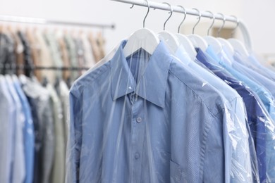 Photo of Dry-cleaning service. Many different clothes in plastic bags hanging on rack indoors, closeup