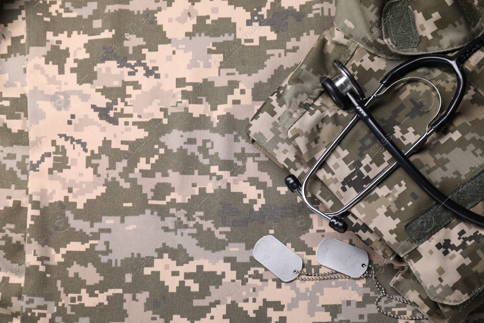 Photo of Stethoscope, military ID tags and soldier uniform on camouflage fabric, flat lay. Space for text
