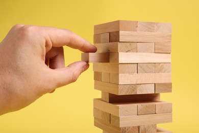 Playing Jenga. Man removing wooden block from tower on yellow background, closeup
