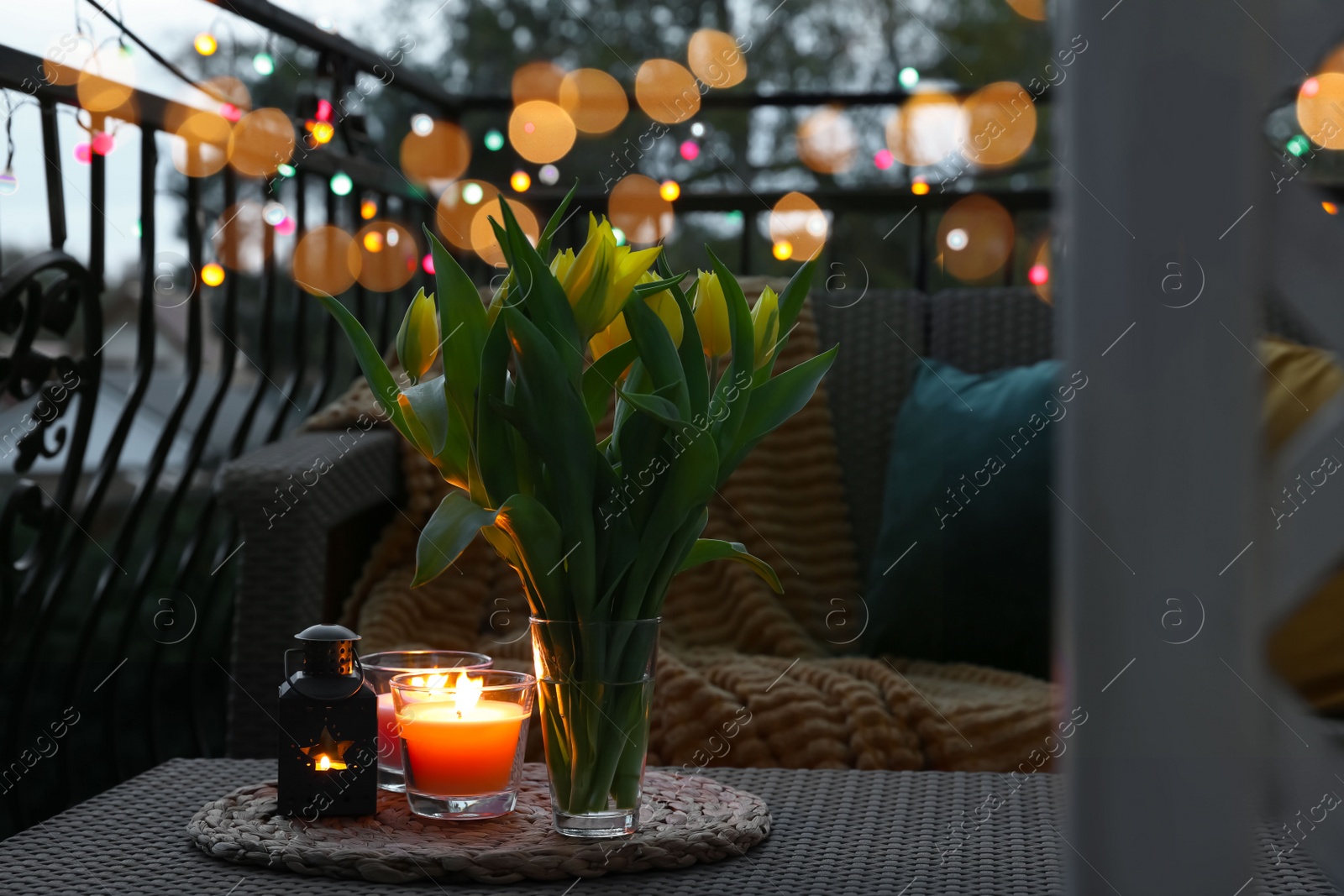 Photo of Soft pillows, blanket, burning candles and yellow tulips on rattan garden furniture in evening