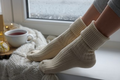 Photo of Woman in warm socks relaxing on window sill at home, closeup