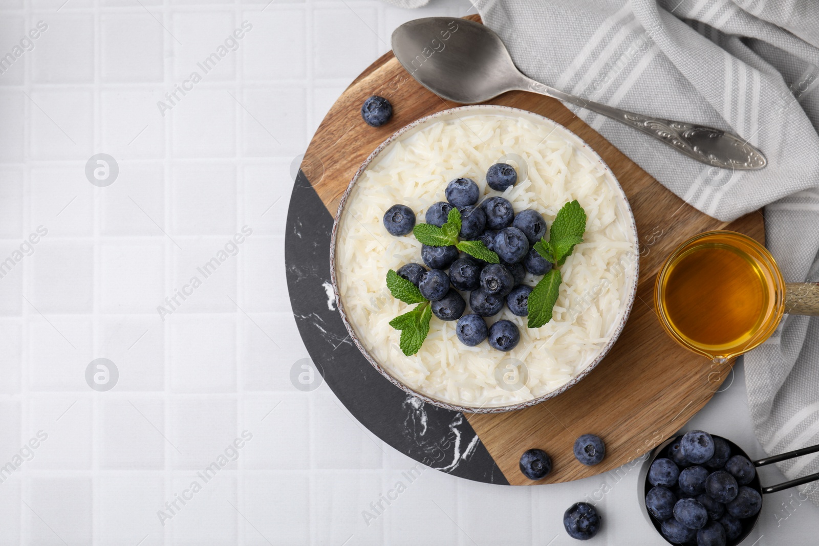 Photo of Bowl of delicious rice porridge with blueberries and mint served on white tiled table, flat lay. Space for text