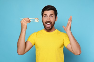 Photo of Emotional man holding sushi roll with chopsticks on light blue background