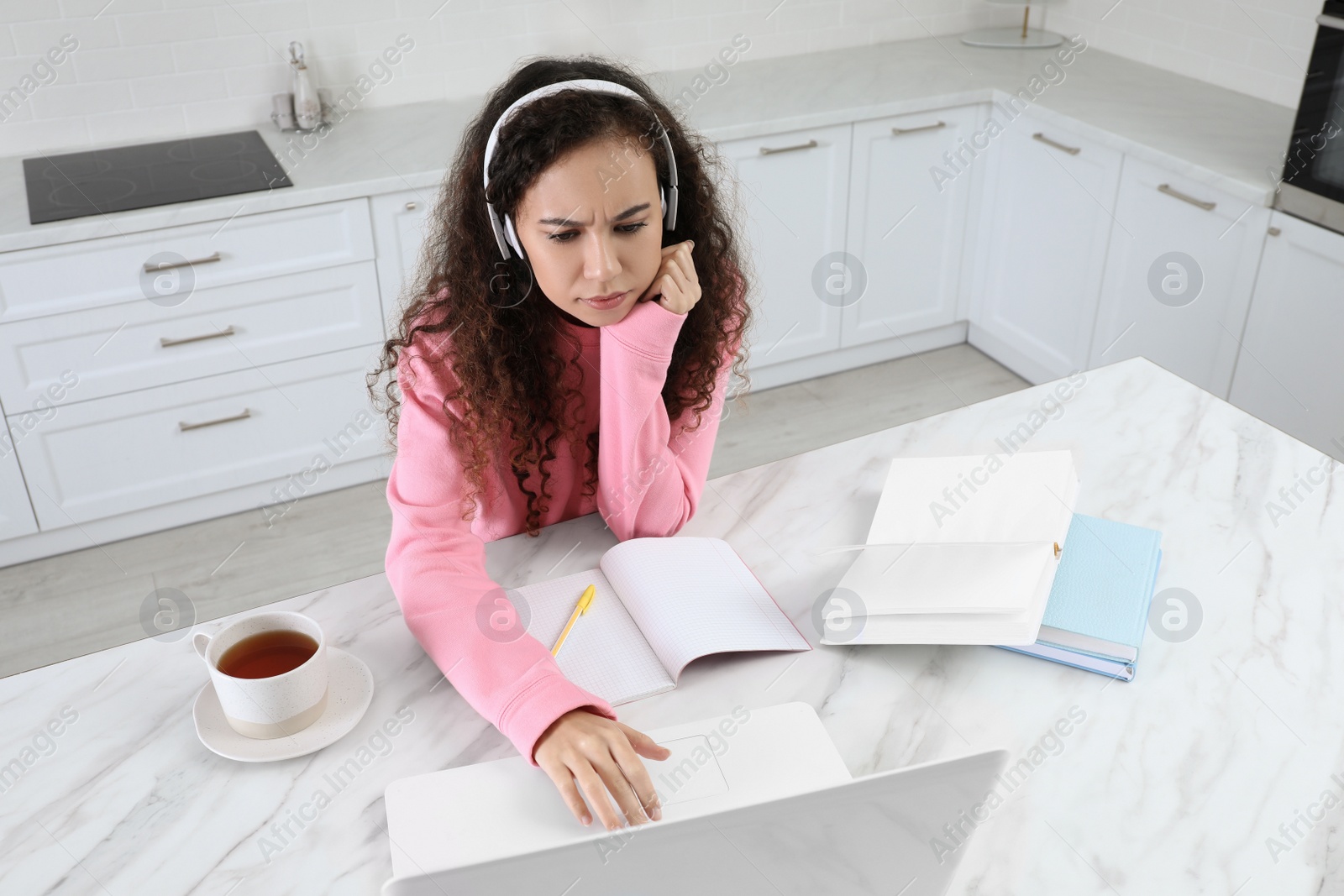 Photo of African American woman with modern laptop and headphones studying in kitchen. Distance learning