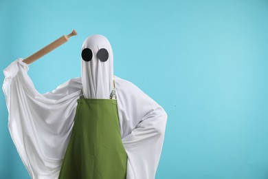 Woman in ghost costume and apron with rolling pin on light blue background, space for text