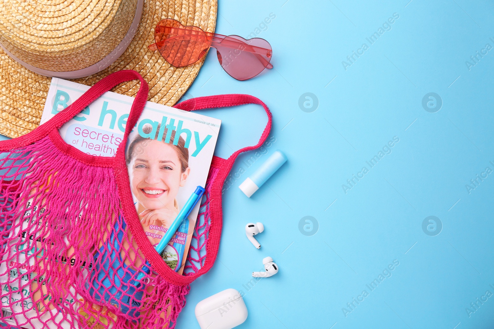 Photo of String bag with magazine, earphones and summer accessories on light blue background, flat lay. Space for text